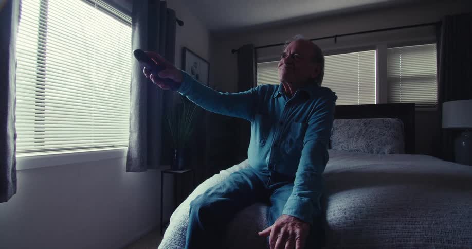 Elderly man sitting on bed and using a television remote control