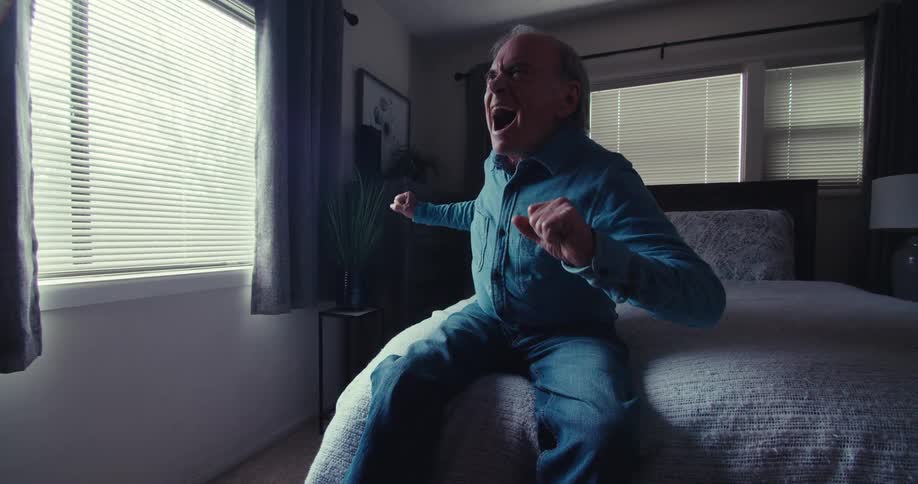 An elderly man wearing blue clothes is sitting on a bed and looking forward while talking and making bothered gestures