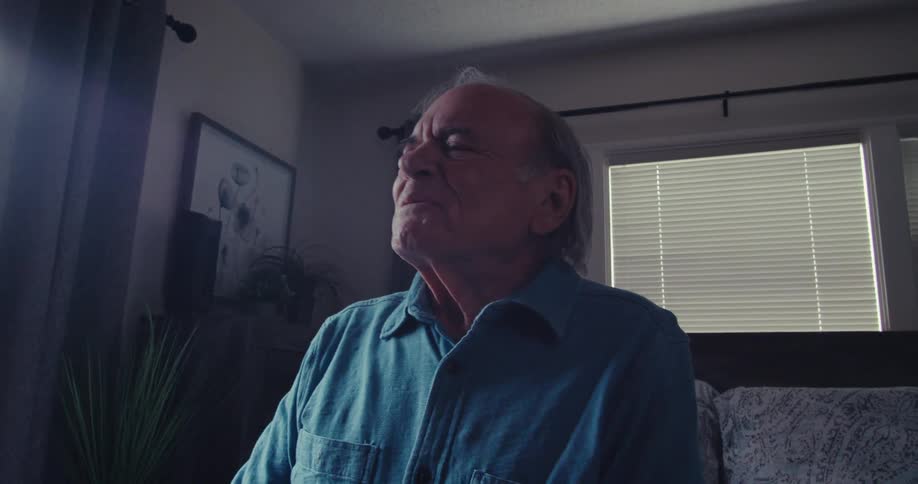 Elderly man sitting on a bed in a bedroom and holding a television remote control