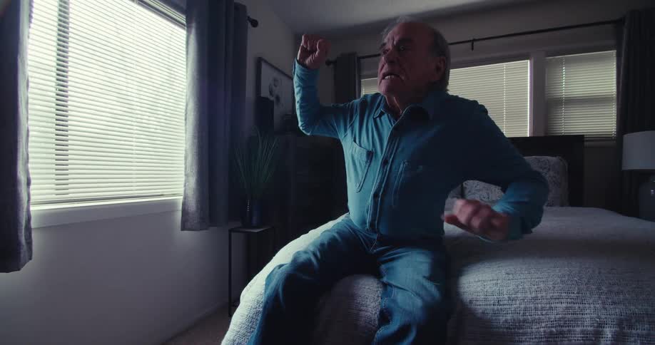 An elderly man sits on his bed in a cozy bedroom at home joyfully celebrating while watching television