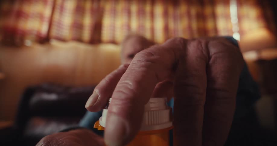 A closeup shows an elderly man in a dimly lit living room unscrewing a pill bottle and taking pills into his hand