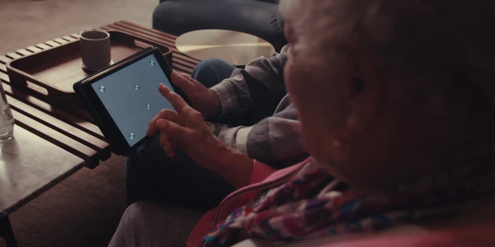 An elderly man and woman sit on a couch looking down at a tablet in the living room