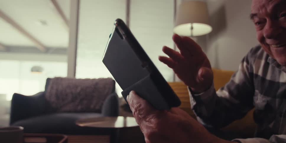 An elderly man and woman are smiling while sitting on the couch looking at a tablet