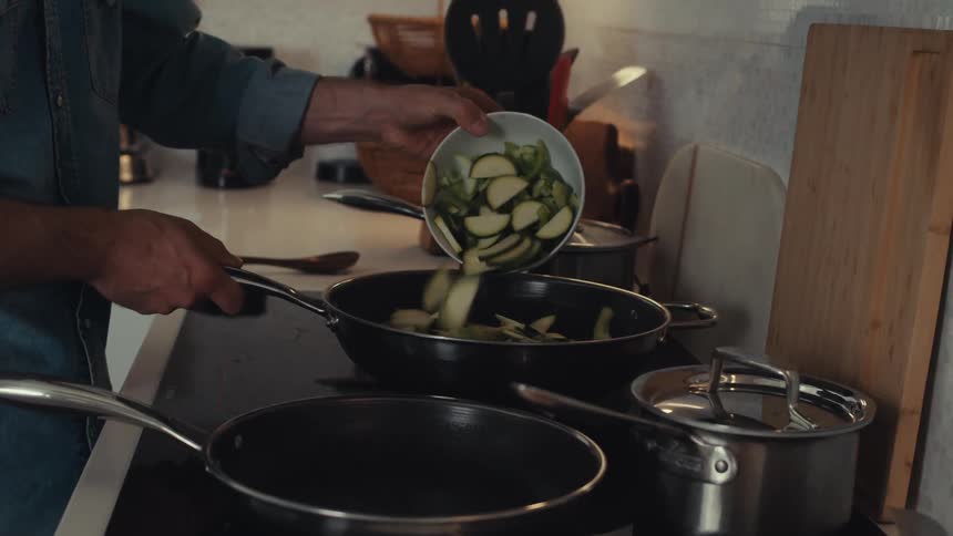 A man is preparing a meal adding chopped zucchini to a pan alongside other ingredients on the kitchen counter