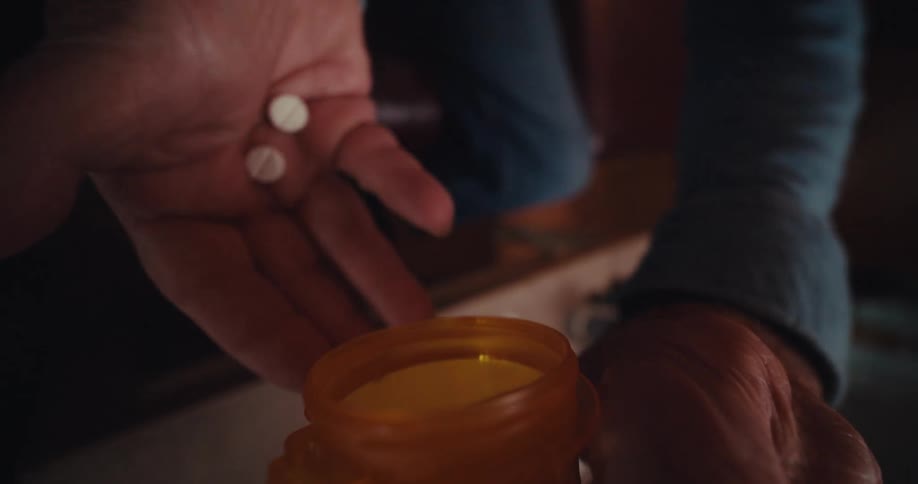 A closeup shows an elderly man in a dimly lit living room unscrewing a pill bottle taking pills into his hand and screwing the bottle closed again