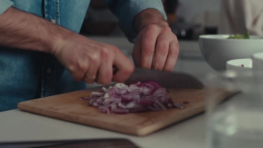 A man in a denim shirt chopping red onions on a wooden cutting board with a knife
