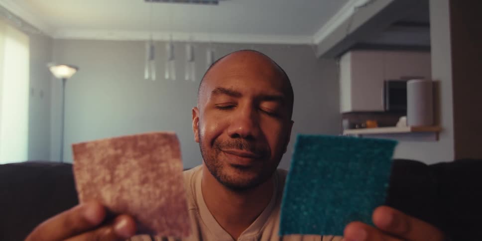 A man sits in a room holding two rectangular objects likely fabric samples as he examines and compares them