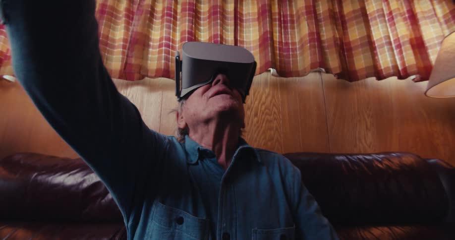 In a cozy living room an elderly man wearing a vr headset points his controller overhead and looks up while moving around as the camera occasionally zooms in and out capturing his immersive interaction