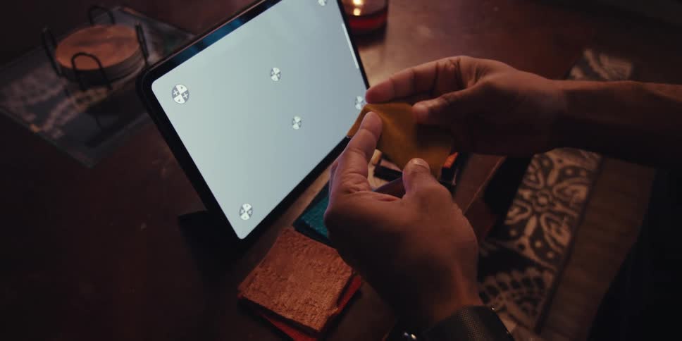 A persons hand picking up some pieces of orange cloth and in front of it there is a tablet with a white screen