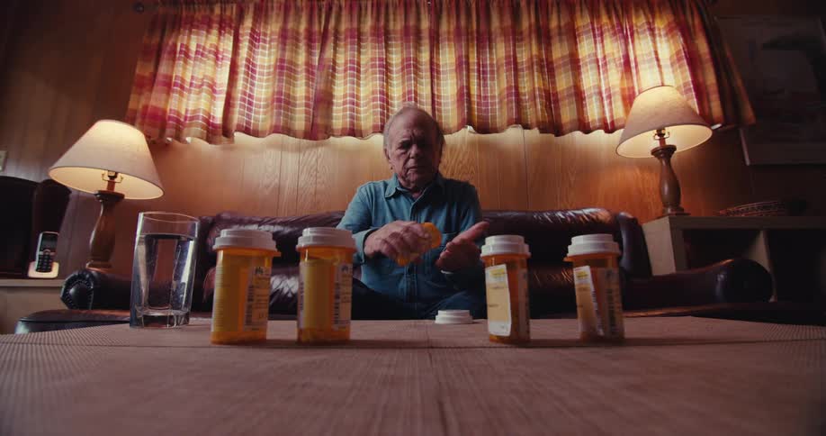 Elderly man sitting in front of several medications and taking a pill and a glass cup next to it