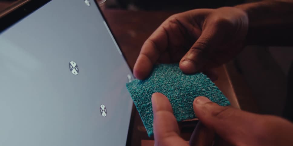 A mans hands holding a piece of fabric next to a screen possibly comparing colors or textures