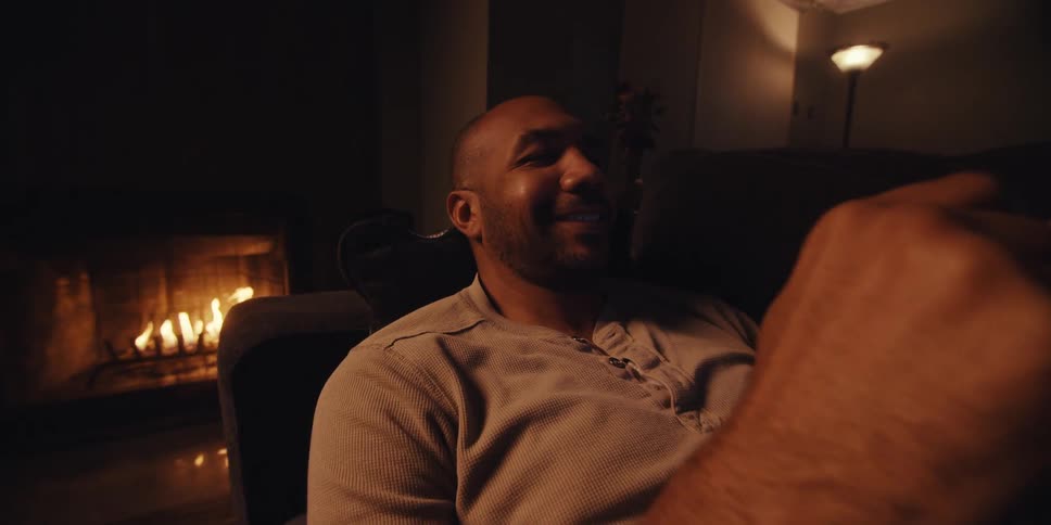 A man relaxes on a couch in a dimly lit room looking at his phone near a cozy fireplace
