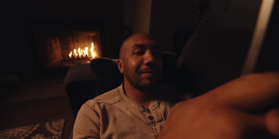A man with a shaved head short beard and lightcolored sweatshirt sits in a dimly lit room illuminated by the warm glow of a fireplace looking at his smartphone with a focused expression