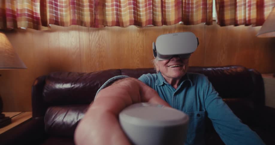 An elderly man sits on a couch in a room fully engaged in playing a virtual reality video game