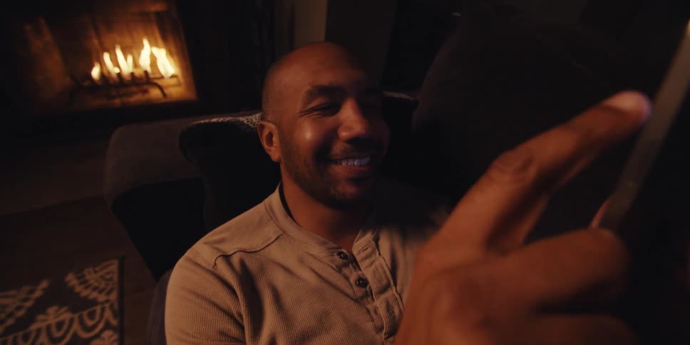 Sitting in a dimly lit room warmed by a fireplaces glow a man with a shaved head short beard and lightcolored sweatshirt looks at his smartphone smiling