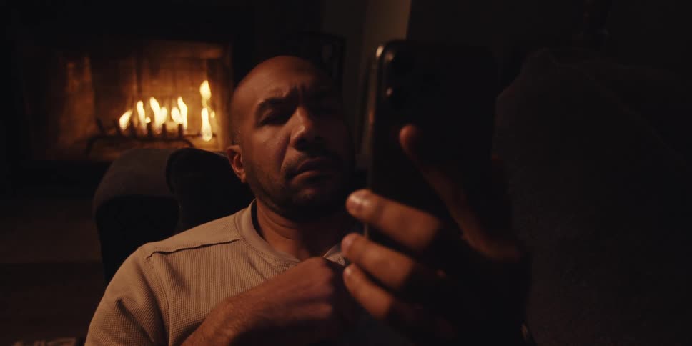 The face of a man who is thinking and then moves the screen of his cell phone and he is sitting on the sofa and behind there is a fireplace