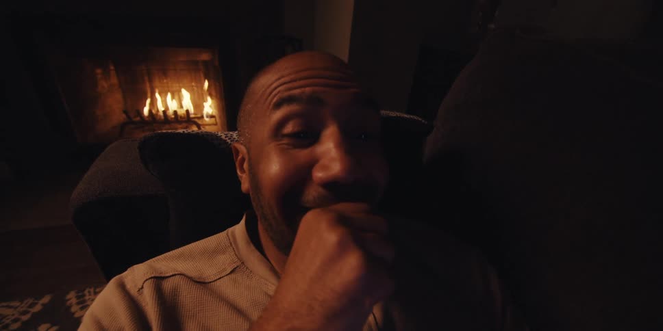 Man reclining on the couch near a burning fire smiles while viewing his smartphone screen