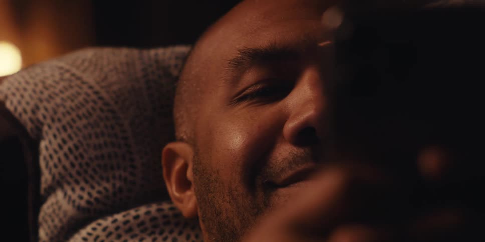 A cheerful bald man in a light colored sweatshirt lies on a couch near the fireplace using his mobile phone