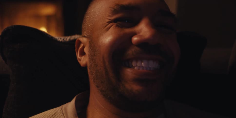 A man lying on a couch in a warmly lit living room celebrates with a fist pump while holding a phone in one hand illuminated by the flickering light of a nearby fireplace