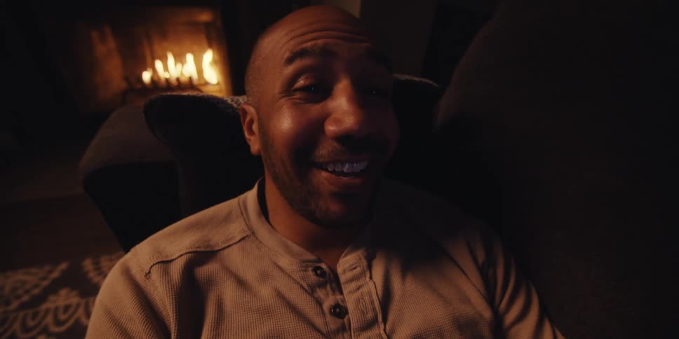 A man lying on the couch next to a fireplace is talking with someone on the smart phone