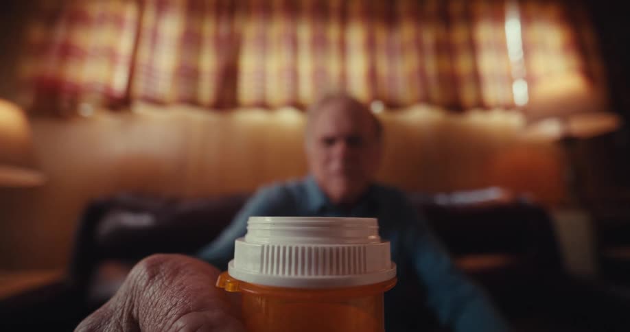 The elderly man is looking at his pill bottles and picks one up for a closer look then chooses the other one to open and take some pills out