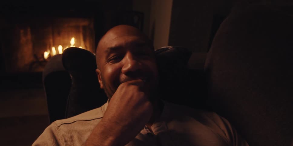 A closeup of a man resting on a couch smiling as he holds a phone in his hand with a dimly lit fireplace in the background