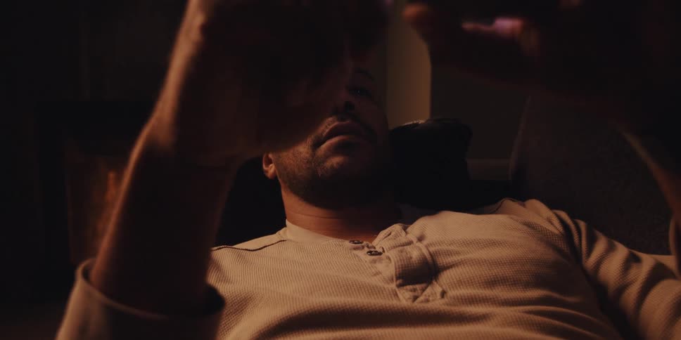 Sitting in a dimly lit room warmed by a fireplaces glow a man with a shaved head short beard and lightcolored sweatshirt looks at his smartphone with a focused expression