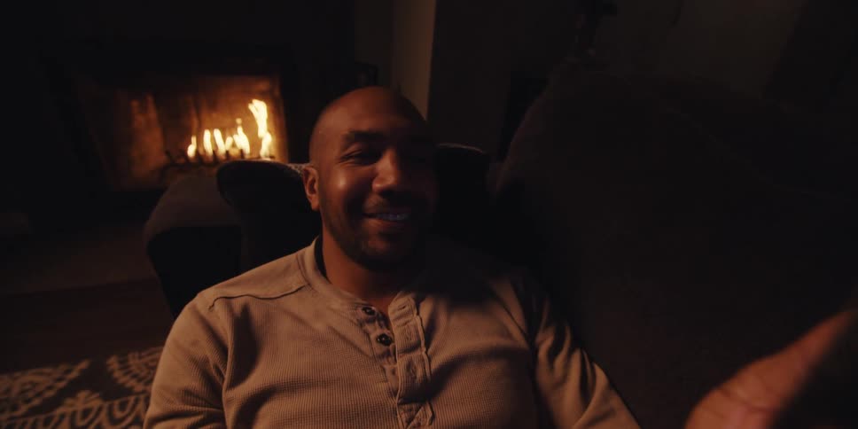 A cheerful bald man wearing a light colored sweatshirt is lying on a couch near the fireplace and using his mobile phone