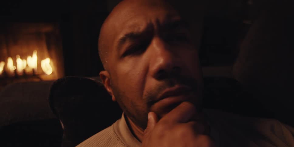 A pensive bald man sits in a dimly lit room with a fireplace lost in thought as he rests his chin on his hand