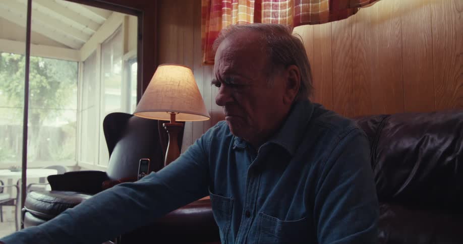 An elderly man sits on a living room couch unscrewing a pill bottle taking a pill and drinking water from a glass