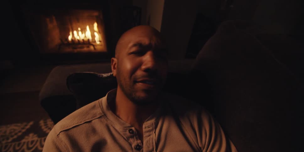 A man sits in a dimly lit room with a fireplace and appears to be having an emotional phone call