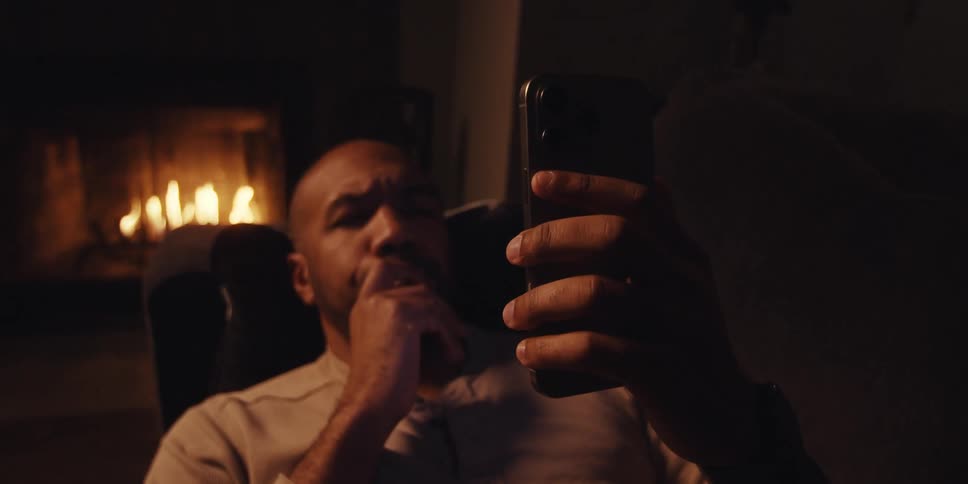 Man lying on the couch concentrates on his smartphone while a fire burns in the fireplace