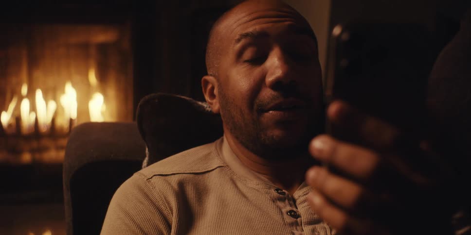 A bald man wearing a light colored shirt is reclined on a couch engaged in a video call on his mobile phone with a fireplace visible in the background
