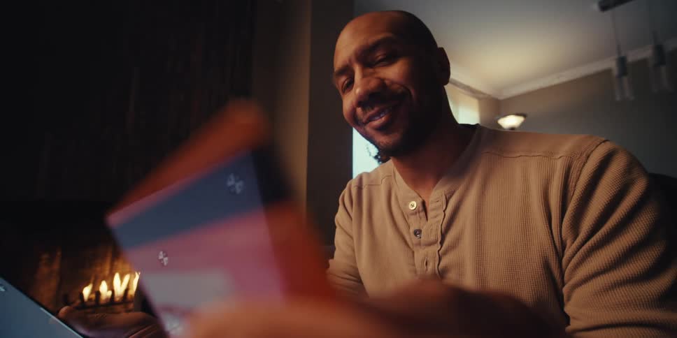 A man sits near a fireplace in a dimly lit room making a payment on a tablet with his credit card