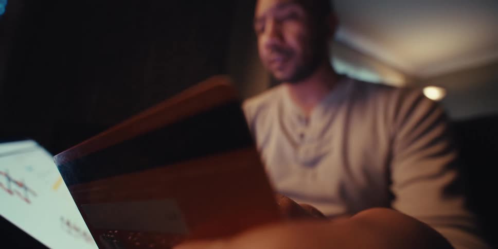 A closeup shows a mans face in a dimly lit room holding a tablet and a credit card