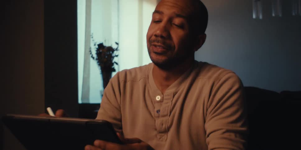A man sits on a couch in a dimly lit room near a fireplace intently focused on a drawing or writing tablet in his hands