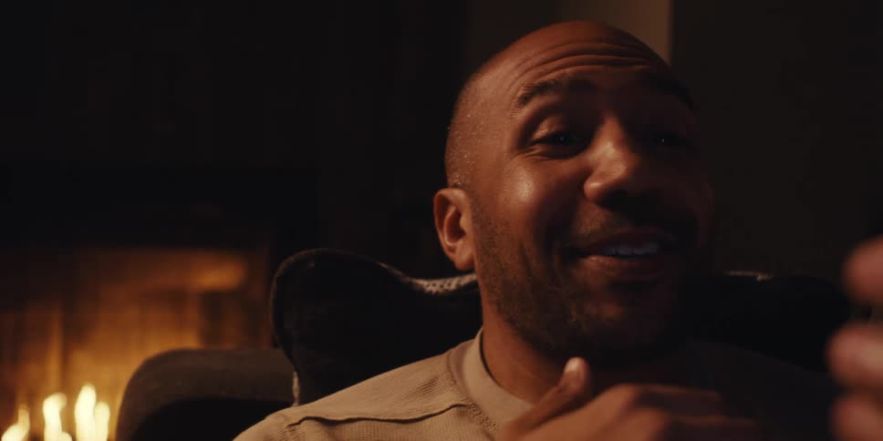 A man sits on a couch in a dimly lit room talking on a video call with a surprised smile as a fireplace flickers softly in the background