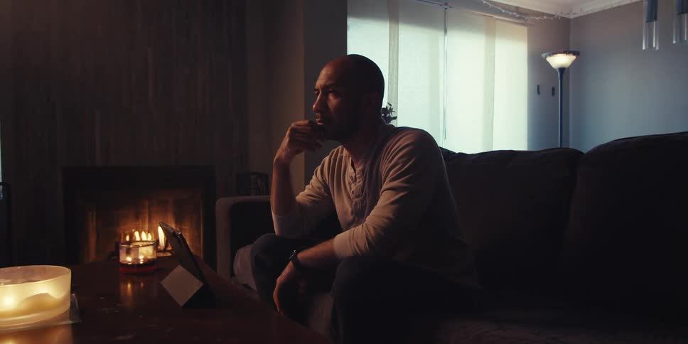 A man with a shaved head and a light colored shirt sits on a couch in a dimly lit room looking down at a tablet on a coffee table next to a fireplace with lit candles