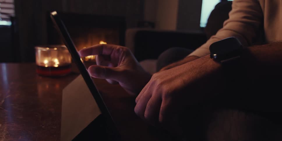 A persons hand using their fingers to scroll through the screen of a tablet that is resting on a table in a dark room with a fireplace in the background