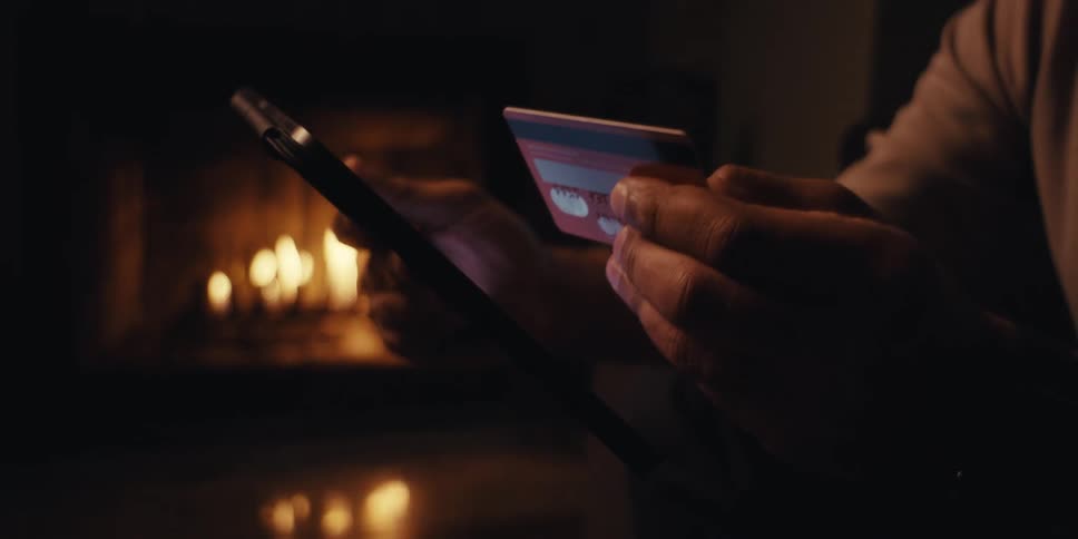 Man holds a credit card while shopping on his tablet in a room warmed by a fireplace