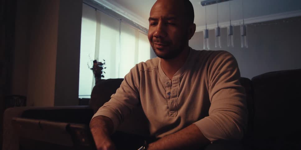 A man is sitting on a couch in a dimly lit room looking at a tablet and smiling