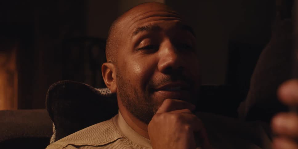 Man with a shaved head and a short beard looking thoughtfully at smartphone having a video call with a warm light source illuminating his face