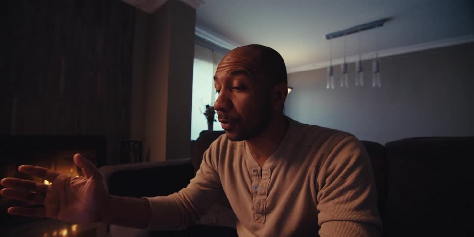 A seated man is talking and doing gestures with his hands in a living room and there is a fireplace in his right side