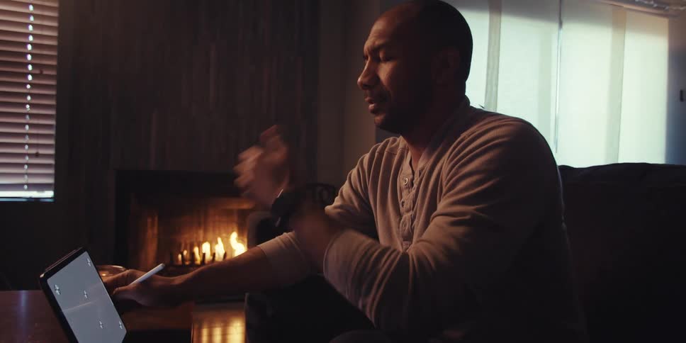A man sits on a couch in a dimly lit room near a fireplace intently focused on a drawing or writing tablet in his hands