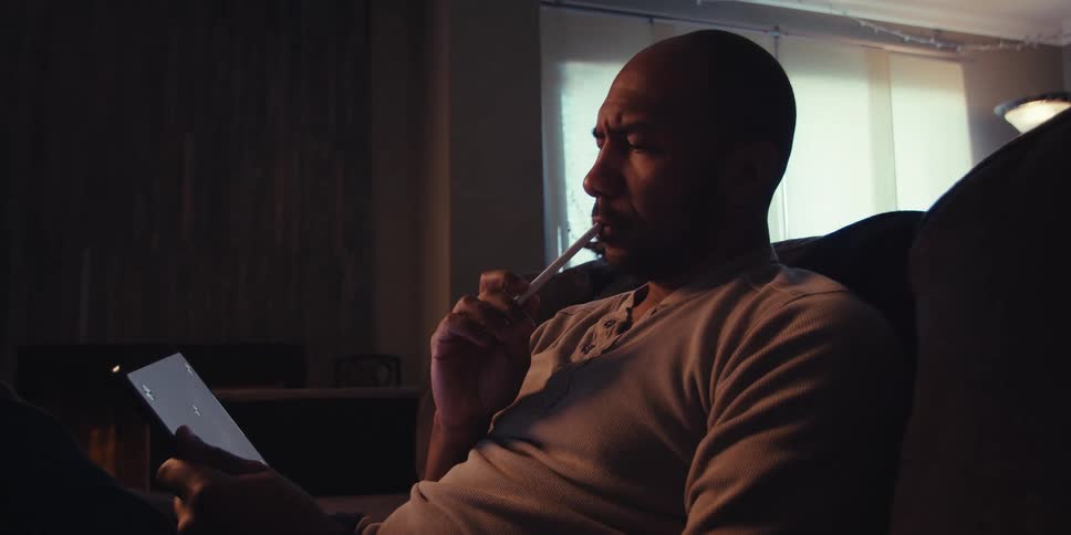 A man sits on a couch in a dimly lit room deeply focused on a drawing or writing tablet in his hands