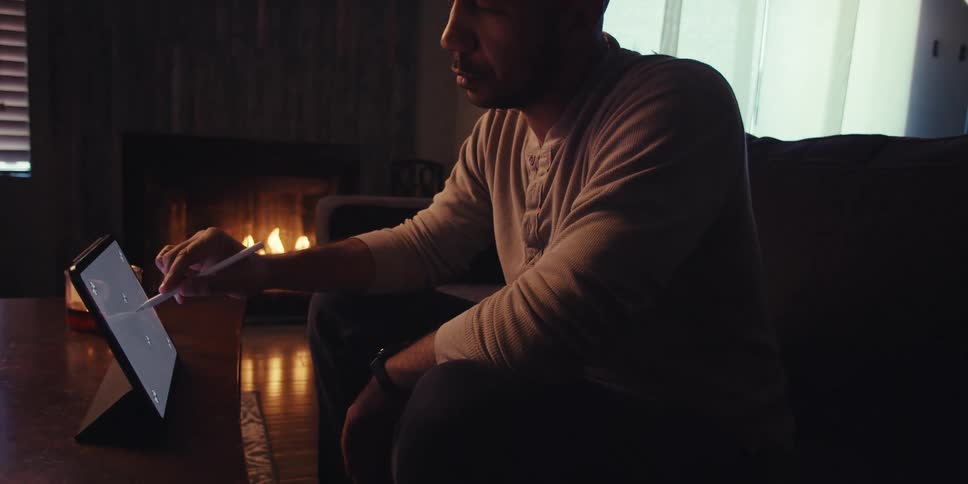 A man sits in a dimly lit room by a fireplace and uses a stylus on a tablet