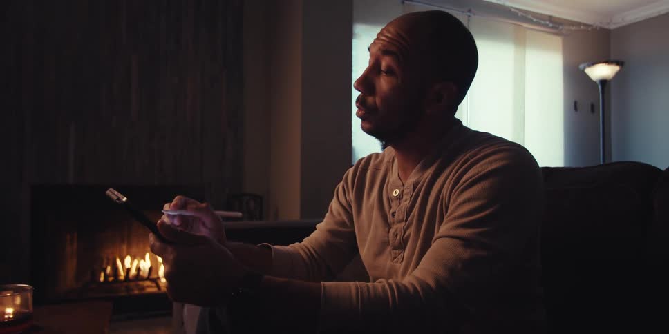 A middle aged man with a shaved head and a short beard sitting in a dimly lit room wearing a beige henley shirt holding a tablet and a stylus