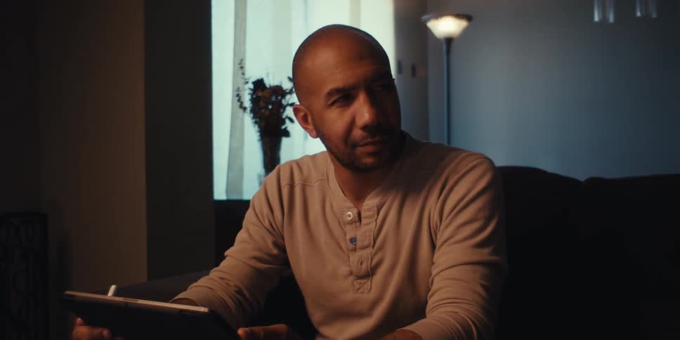 A man with a shaved head and a light colored shirt sits on a couch in a dimly lit room looking down at a tablet and thinking