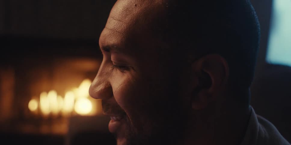 The face of a man who is sitting in a partially dark room and there is a fireplace behind him and he begins to smile