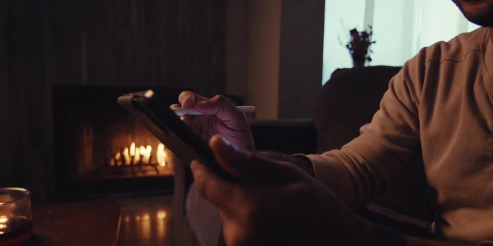 Man sitting in a living room containing a lit fire smiles while using a stylus on a tablet